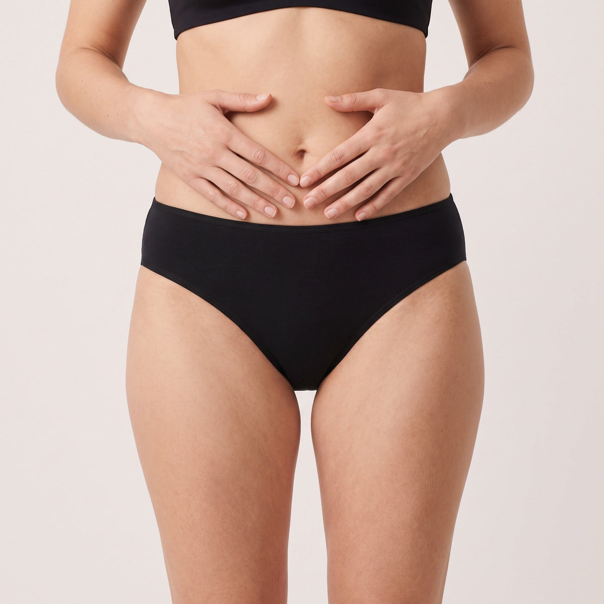 Culotte bikini classique Louma noire portée, vue de face, mains posées sur le ventre